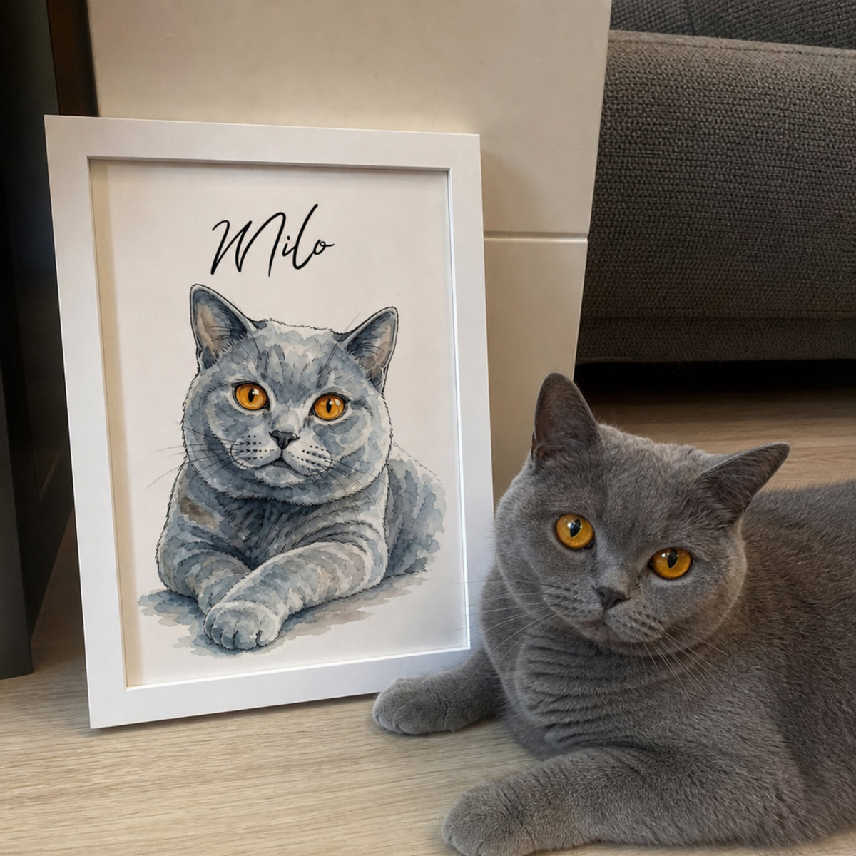 Gray British Shorthair cat lying beside a framed custom watercolor pet portrait personalized with the name Milo.