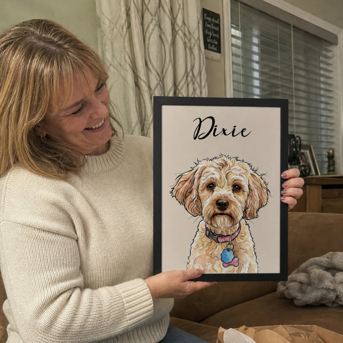 A woman sitting on a couch holds up a framed custom pet portrait featuring a doodle-style illustration of a dog named 'Dixie'. An open cardboard box with tissue paper is visible in the foreground, and a living room setting is in the background.