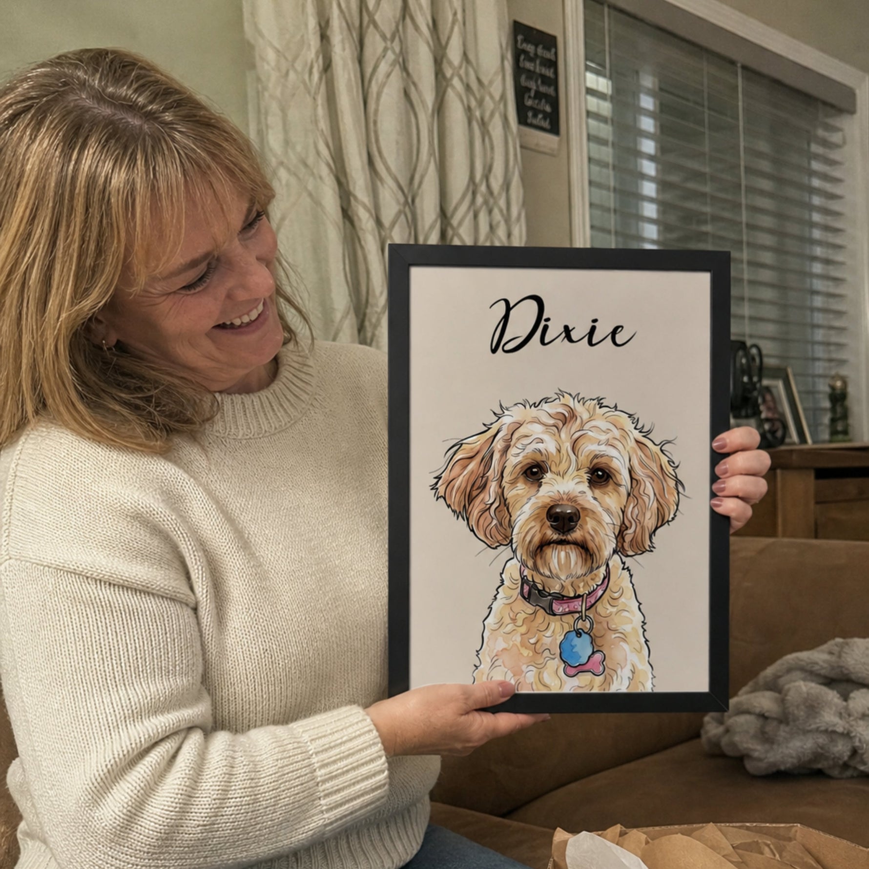 A woman sitting on a couch holds up a framed custom pet portrait featuring a doodle-style illustration of a dog named 'Dixie'. An open cardboard box with tissue paper is visible in the foreground, and a living room setting is in the background.